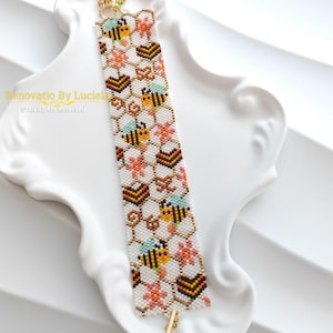 Könnte beinhalten: Ein Perlenarmband mit einem Bienenwabenmuster, Bienen, Blumen und Herzen. Das Armband ist weiß mit gelben, braunen, orangefarbenen und blauen Akzenten. Der Verschluss ist goldfarben. Der Text "Renovatio By Lucielle" ist sichtbar.