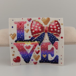 May include: A white canvas with a beaded design of the word "LOVE" in pink, purple, and blue with a red and white polka dot bow in the center. The design is surrounded by small red and gold hearts.