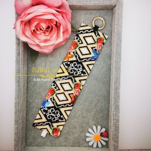 Può includere: Un braccialetto di perline bianco e nero con un motivo geometrico e accenti floreali colorati. Il braccialetto è esposto in una scatola di velluto grigio con una rosa rosa e una margherita bianca.