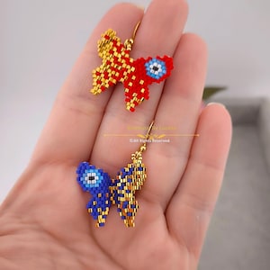 Puede incluir: Un par de pendientes de mariposa dorados con abalorios de ojo turco azul y rojo. Los pendientes están hechos de pequeñas cuentas y tienen un diseño delicado.