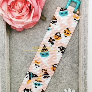 May include: A pink and white beaded bracelet with a Halloween theme. The bracelet features black cats, ghosts, pumpkins, and witch hats. The bracelet has a turquoise clasp.