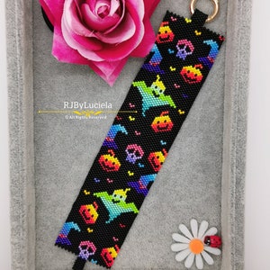 May include: A black beaded bracelet with a colorful pattern of ghosts, pumpkins, and witch hats. The bracelet is in a gray velvet box with a pink rose and a white daisy with a ladybug.