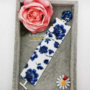 May include: A white and blue beaded bracelet with a floral pattern. The bracelet is in a gray velvet box with a pink rose and a blue flower charm.
