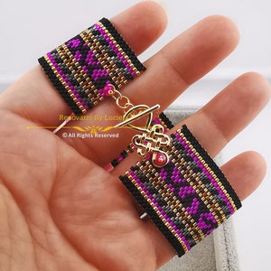 Op de afbeelding: Een zwarte en gouden kralen armband met roze accenten en een gouden sluiting. De armband is gemaakt van kleine kralen en heeft een geometrisch patroon. De sluiting is in de vorm van een knoop.