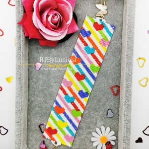 Può includere: Un braccialetto colorato con perline su sfondo bianco e un motivo di strisce diagonali e cuori. Il braccialetto ha una chiusura dorata con un ciondolo a forma di farfalla. Il braccialetto è esposto in una scatola grigia con una rosa rosa e altri elementi decorativi.