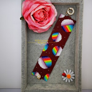 May include: A beaded bracelet with a maroon background and colorful rainbow circles. The bracelet has a gold clasp and is displayed in a gray velvet box with a pink rose and a white daisy.