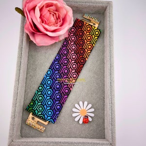 May include: A rainbow coloured beaded bracelet with a geometric pattern. The bracelet is made with small, colourful beads and has a gold clasp.