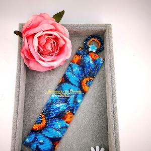 Pode incluir: Um bracelete de contas azul e laranja com um padrão floral. O bracelete é apresentado numa caixa de joias de veludo cinzento com uma rosa cor-de-rosa e uma margarida branca.