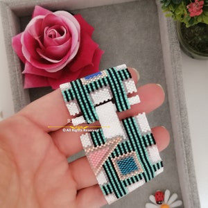 May include: A white, black, green, pink and blue beaded bracelet with a geometric pattern. The bracelet is made of small beads and has a unique design. The bracelet is on a hand and the text "R.Joyhucicela ©All Rights Reserved" is visible.