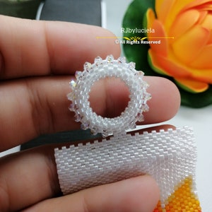 May include: A white beaded ring with clear crystals, held in a hand. The ring is made with small beads and has a circular shape. The background is blurred and shows a flower.