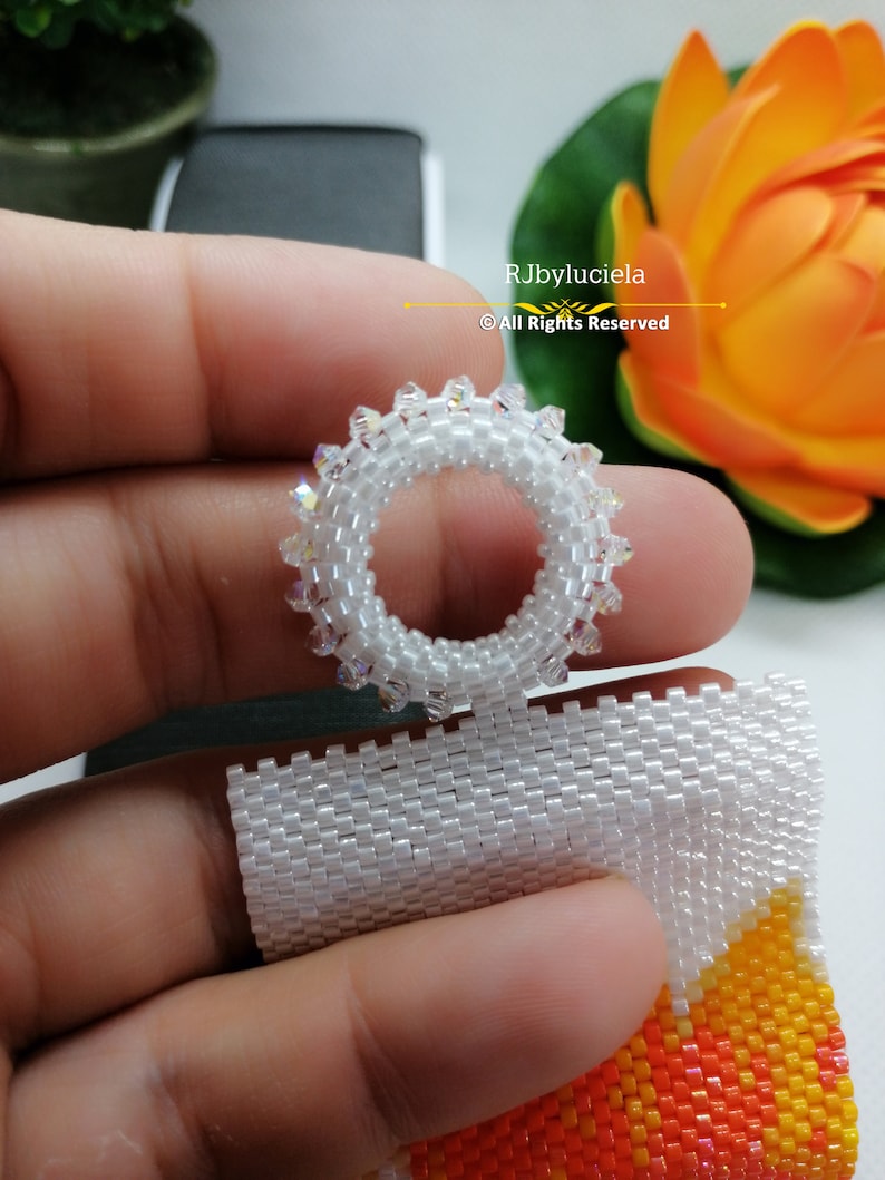 May include: A white beaded ring with clear crystals, held in a hand. The ring is made with small beads and has a circular shape. The background is blurred and shows a flower.