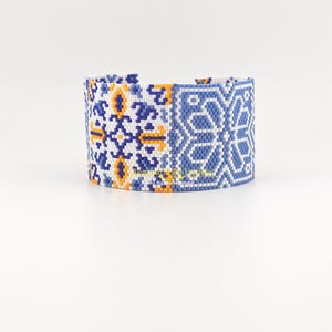Peut inclure: Large bracelet en perles avec un motif géométrique blanc, bleu et jaune. Le bracelet présente un motif répété de formes florales et géométriques stylisées. Le bracelet mesure environ 5 cm de large.