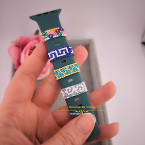 May include: A green silicone Apple Watch band with four colourful beaded rings. The rings feature different patterns, including flowers, geometric shapes, and a blue and white Greek key design. The text "Renovatio By Luciela" and "© All Rights Reserved" is visible on the band.