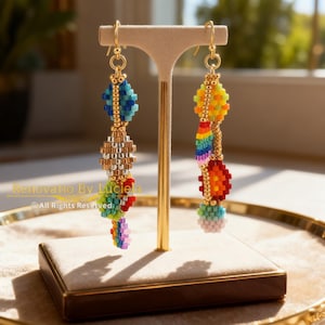 K&ouml;nnte beinhalten: Goldfarbene Perlenohrringe mit Regenbogenfarben. Die Ohrringe haben eine Reihe von farbigen Perlenabschnitten, darunter Blau, Gold, Rot und Gr&uuml;n, mit einem h&auml;ngenden Regenbogen-Design. Die Ohrringe werden auf einem beigen St&auml;nder ausgestellt.