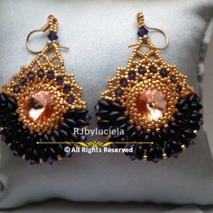 Puede incluir: Un par de pendientes hechos a mano con un diseño de cuentas doradas. Los pendientes presentan una gema central de color melocotón rodeada de cuentas azul oscuro y doradas. Los pendientes se exhiben sobre un cojín azul claro.