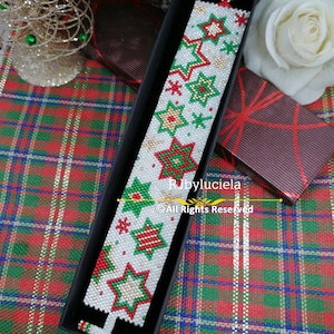 Può includere: Un braccialetto di perline bianche a tema natalizio. Il braccialetto presenta perline rosse, verdi e dorate in un motivo a stella. Il braccialetto è esposto in una scatola nera su una superficie a quadri rossi e verdi.