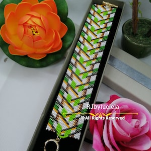 May include: A green, white and gold beaded bracelet with a geometric pattern. The bracelet is in a small box with a black velvet lining. The text "RJbyluciela ©All Rights Reserved" is printed on the box.