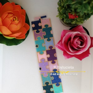 Può includere: Un braccialetto colorato con perline a forma di pezzo di puzzle. Il braccialetto è realizzato con perline blu, verdi, viola, rosa e dorate. Il braccialetto è su una superficie bianca.
