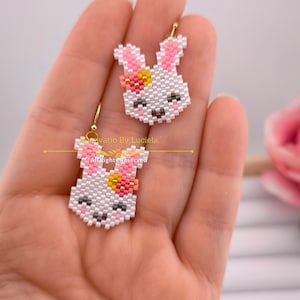 May include: A pair of white bunny earrings with pink ears and a pink flower on the head. The earrings are made of beads and have gold hoops.