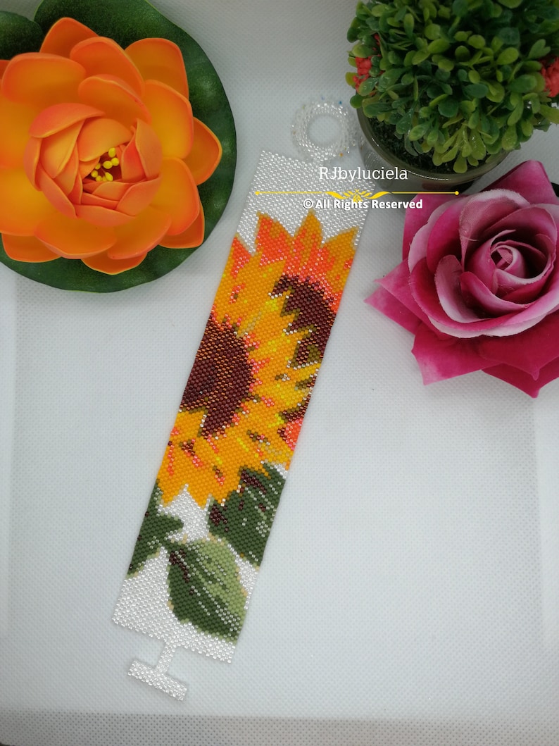 May include: A white beaded bracelet with a colorful sunflower design. The bracelet has a clasp and is made with small beads. The text "RJbyluciela &copy; All Rights Reserved" is printed on the bracelet.
