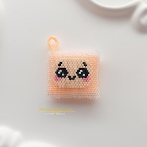 Può includere: Un ciondolo quadrato di perline nei toni del pesca e del bianco con un design centrale raffigurante un volto con occhi neri, una piccola bocca nera e guance rosa. Il ciondolo è realizzato con piccole perline uniformi e include un occhiello color pesca.