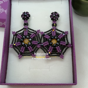 Peut inclure: Une paire de boucles d'oreilles en forme de toile d'araignée en perles violettes et noires dans une boîte cadeau violette. Les boucles d'oreilles sont pendantes et présentent un motif de toile d'araignée noire et violette avec un centre jaune.