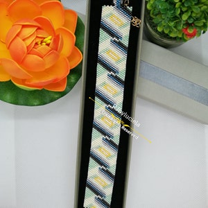 May include: A white, black, blue and yellow beaded bracelet with a gold clasp. The bracelet is in a small, grey box with a black interior.