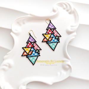 May include: A pair of colourful beaded earrings with a geometric design. The earrings feature multiple stacked triangles in various colours, including purple, yellow, pink, and blue, outlined in black. Gold-coloured ear wires are attached. The earrings are displayed on a white, decorative plate.