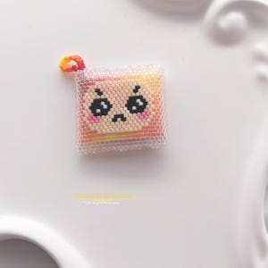 May include: A square-shaped beaded charm featuring a gradient of yellow, orange, and pink colours. The charm displays a face with black eyes, a small mouth, and pink cheeks, all encased in a layer of clear, translucent beads with a small loop.