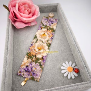 May include: A beaded bracelet featuring a floral design with white roses and purple flowers. The bracelet is made with seed beads and has a gold clasp.