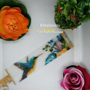May include: A white beaded bracelet with a colorful hummingbird and floral design. The bracelet has a gold clasp and is made with seed beads. The text "RJbyluciela &copy; All Rights Reserved" is printed on the bracelet.