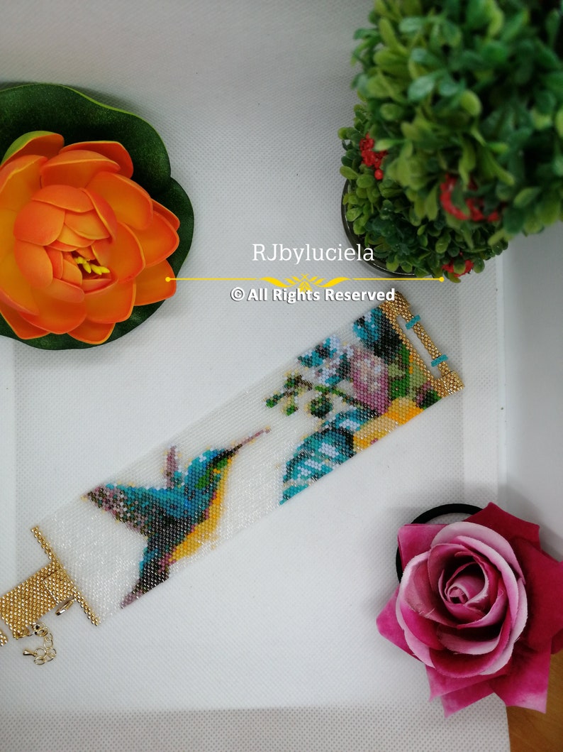 May include: A white beaded bracelet with a colorful hummingbird and floral design. The bracelet has a gold clasp and is made with seed beads. The text "RJbyluciela &copy; All Rights Reserved" is printed on the bracelet.
