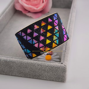 May include: A black beaded bracelet with a geometric pattern of colorful triangles. The bracelet is in a gray velvet box with a white flower.