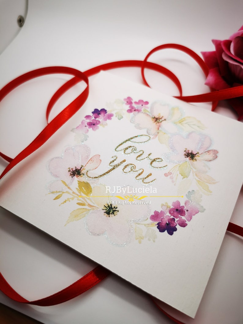 May include: A white card with a watercolor floral wreath and the words "love you" in gold glitter lettering. The card is signed "RJByLuciela" and includes the copyright notice "&copy;All Rights Reserved".