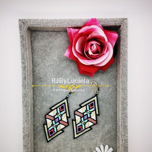 Puede incluir: Un par de pendientes de cuentas geom&eacute;tricas en una caja de terciopelo gris con una rosa rosa y una mariquita en una margarita. Los pendientes son negros, blancos, rosas, azules y dorados.