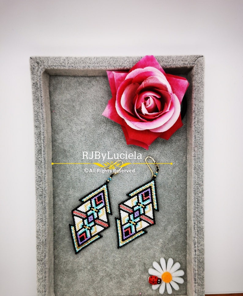 Puede incluir: Un par de pendientes de cuentas geom&eacute;tricas en una caja de terciopelo gris con una rosa rosa y una mariquita en una margarita. Los pendientes son negros, blancos, rosas, azules y dorados.