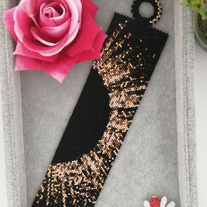 Può includere: Un braccialetto di perline nere e dorate con un motivo geometrico. Il braccialetto ha una chiusura rotonda ed è realizzato con perline piccole.