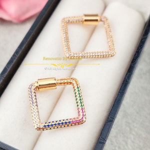 May include: Two gold square-shaped charms with a clear crystal design. One charm has a rainbow of colored crystals around the edge. The other charm has clear crystals around the edge. The charms are in a black box with the text "Renovatio By Lorena" and "© All Rights Reserved".