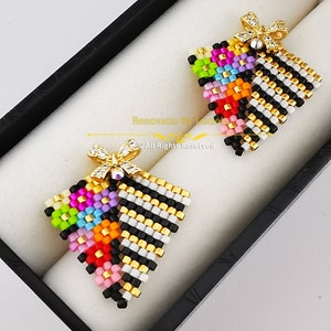 Puede incluir: Un par de pendientes triangulares de colores con rayas blancas y negras y un dije de lazo dorado. Los pendientes se muestran en una caja negra.