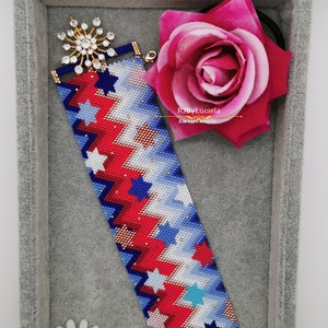 May include: A beaded bracelet with a red, white, and blue star pattern. The bracelet is in a gray velvet box with a pink rose and a small white daisy.