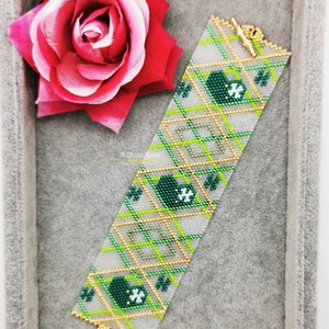 May include: A green and gold beaded bracelet with a clover design. The bracelet is in a grey velvet box with a pink rose and a white daisy with a red ladybug.