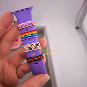 May include: A purple Apple Watch band with four colourful beaded rings. The rings are rainbow, brown and white, and green and red.