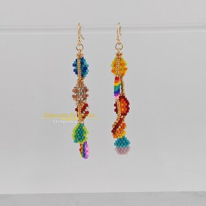 K&ouml;nnte beinhalten: Ein Paar Perlenohrringe mit goldfarbenen Haken. Die Ohrringe weisen farbenfrohe Perlenmuster in Blau-, Gr&uuml;n-, Braun-, Gelb-, Orange-, Rot- und Rosat&ouml;nen auf. Die Ohrringe h&auml;ngen vor einem hellen Hintergrund.