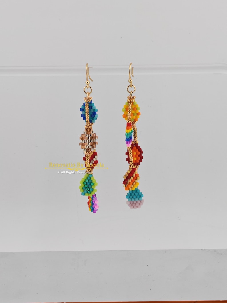 K&ouml;nnte beinhalten: Ein Paar Perlenohrringe mit goldfarbenen Haken. Die Ohrringe weisen farbenfrohe Perlenmuster in Blau-, Gr&uuml;n-, Braun-, Gelb-, Orange-, Rot- und Rosat&ouml;nen auf. Die Ohrringe h&auml;ngen vor einem hellen Hintergrund.