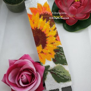 May include: A beaded bracelet with a colorful sunflower design. The bracelet is white with a green leaf and a brown center. The text "RJbyluciela © All Rights Reserved" is on the bracelet.