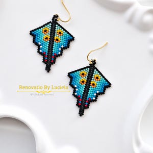 May include: A pair of beaded earrings featuring a geometric kite shape with a gradient of blue, black, and yellow beads, accented with small red details and gold-toned hooks. The text Renovatio By Luciela All Rights Reserved is visible.