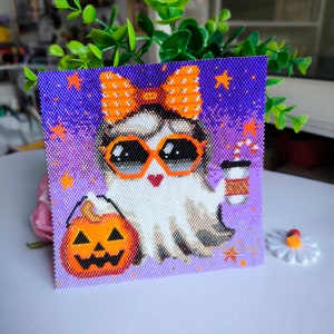 May include: A purple and white beaded ghost with orange and white bow, sunglasses, and a pumpkin bucket. The ghost is holding a cup of coffee with a candy cane stirrer. The background is a purple gradient with white stars.