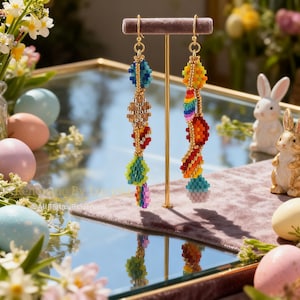 K&ouml;nnte beinhalten: Bunte Perlenohrringe mit goldfarbenen Haken. Die Ohrringe weisen eine Vielzahl von Formen und Farben auf, darunter Blau, Gold, Rot, Gr&uuml;n und Regenbogenfarben. Sie werden auf einem Samtst&auml;nder pr&auml;sentiert, mit Ostereiern und Hasenfiguren im Hintergrund.