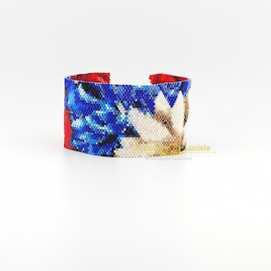 May include: A wide cuff bracelet with a floral design. The bracelet features a red interior and a blue, white, and tan exterior. The beaded design is intricate and detailed, showcasing a flower pattern. The bracelet is a statement piece.
