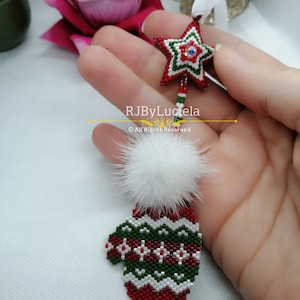 Könnte beinhalten: Ein roter und grüner Weihnachtsbaumschmuck aus Perlen in Form einer Mütze mit einem weißen Bommel oben. Ein Stern aus Perlen mit einem blauen Strassstein in der Mitte hängt über der Mütze. Der Schmuck wird in einer Hand gehalten. RJByLuciela © Alle Rechte vorbehalten.